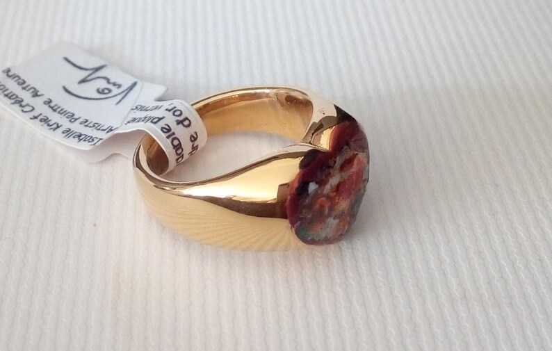 May include: A gold-colored ring with a rounded band and a colorful, abstract design on the face. The ring has a smooth, polished finish. A white tag with text is attached to the ring.