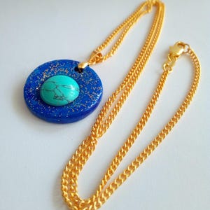 May include: A gold-colored chain necklace with a pendant. The pendant is a blue circle with gold flecks and a turquoise stone in the center. The necklace is displayed on a white surface.