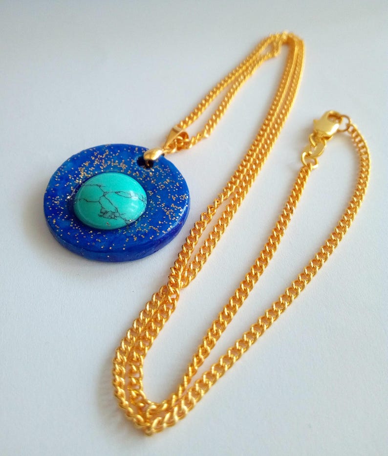 May include: A gold-colored chain necklace with a pendant. The pendant is a blue circle with gold flecks and a turquoise stone in the center. The necklace is displayed on a white surface.
