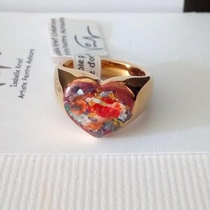 May include: A gold-colored ring with a heart-shaped centerpiece. The heart is filled with a colorful, abstract design featuring red, orange, and white hues. The ring is set against a white background.