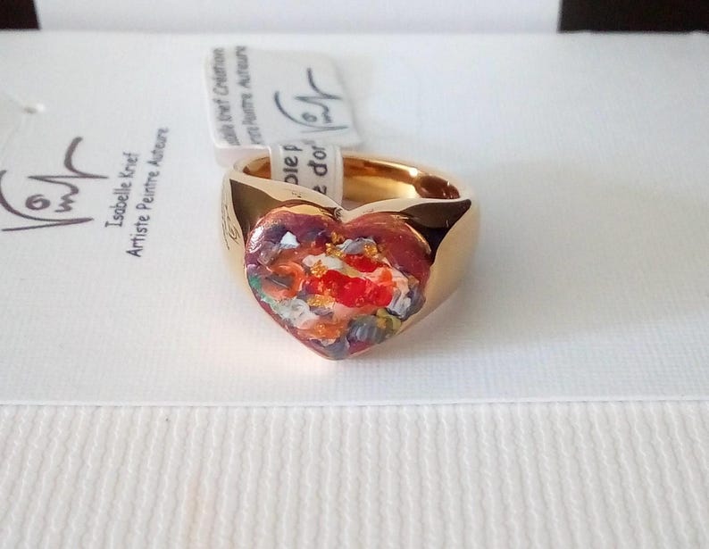 May include: A gold-colored ring with a heart-shaped centerpiece. The heart is filled with a colorful, abstract design featuring red, orange, and white hues. The ring is set against a white background.