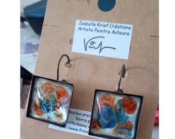 May include: A pair of square earrings with bronze-toned metal frames and hook wires. The centers feature a glossy, abstract design with swirls of orange, blue, and white paint. The earrings are attached to a brown cardboard display card with text.
