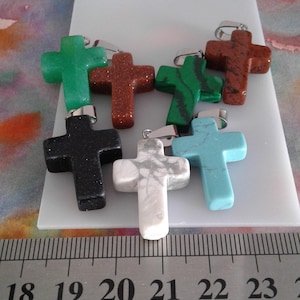 以下が含まれることがあります： 緑、茶色、黒、白、ターコイズなど、さまざまな色の石で作られた7つのクロス型ペンダントのコレクション。各クロスには、チェーンに取り付けるためのシルバー色のベイルが付いています。クロスは白い表面に表示されています。