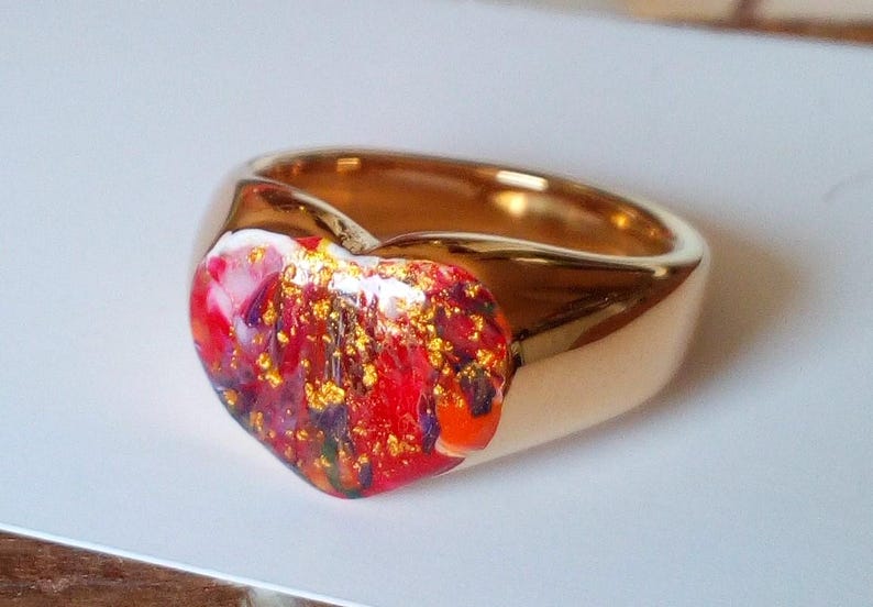 May include: A gold-colored ring with a heart-shaped centerpiece. The heart is filled with a mix of red, orange, purple, and gold-colored flecks. The ring has a smooth, polished finish.