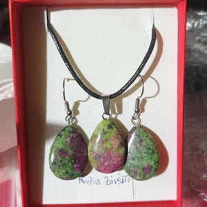 Peut inclure: Un ensemble collier et boucles d'oreilles avec des pierres de rubis zoisite en forme de goutte. Les pierres présentent des couleurs vertes, roses et violettes. Le collier a un cordon noir et les boucles d'oreilles des crochets argentés. L'ensemble est présenté dans une boîte rouge.