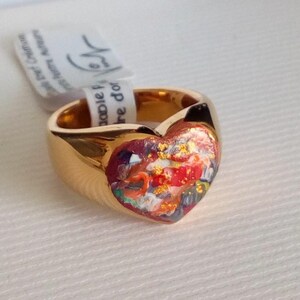 May include: A gold-colored ring with a heart-shaped face. The heart is filled with a colorful, abstract design featuring red, orange, and blue hues. A white tag is attached to the ring.