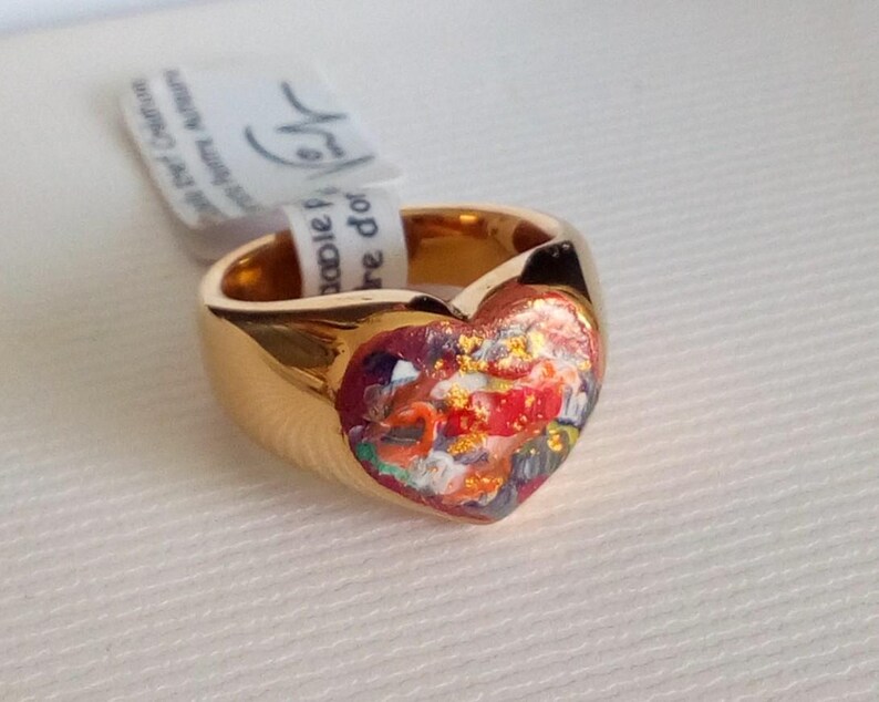 May include: A gold-colored ring with a heart-shaped face. The heart is filled with a colorful, abstract design featuring red, orange, and blue hues. A white tag is attached to the ring.