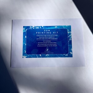 以下が含まれることがあります： 「SUN PRINTING KIT」と書かれた青い長方形が中央にある白いカード。 キットには、感光紙と自然にインスパイアされたアートを作成するためのガイドが含まれています。 水と想像力で創造性を促します。