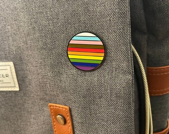 May include: A round enamel pin with a rainbow stripe design. The pin is on a gray fabric background.