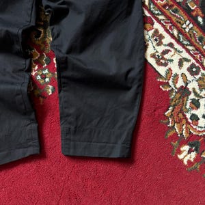 May include: Close-up of black trousers with a slightly textured appearance. The trousers are folded at the bottom hem. The background is a red rug with a detailed pattern in various colours.
