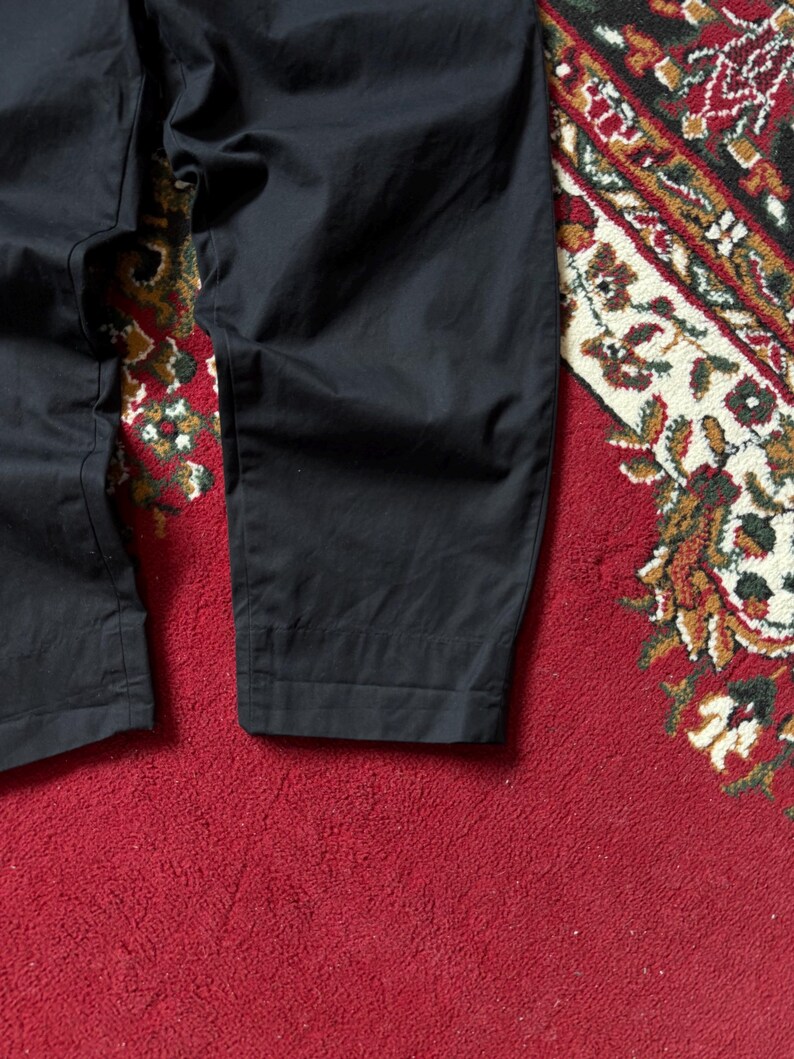 May include: Close-up of black trousers with a slightly textured appearance. The trousers are folded at the bottom hem. The background is a red rug with a detailed pattern in various colours.