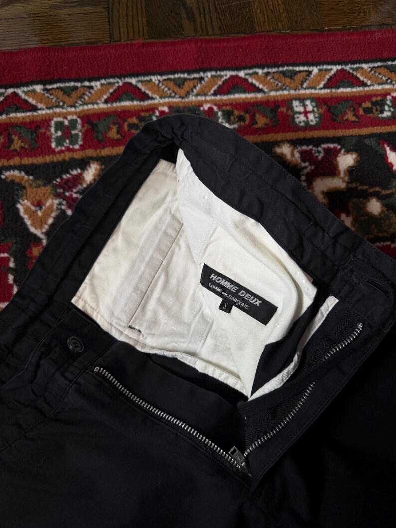 May include: Black trousers with a white lining visible at the waist. The trousers feature a zip and button fastening. The label inside reads "HOMME DEUX COMME des GAR&Ccedil;ONS". Size S.