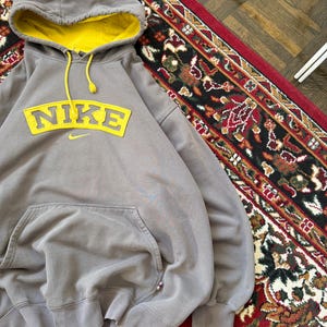 May include: A gray Nike hoodie with a yellow interior hood and yellow drawstring. The Nike logo is prominently displayed in yellow across the chest. The hoodie has a front pocket and is displayed on a red rug.