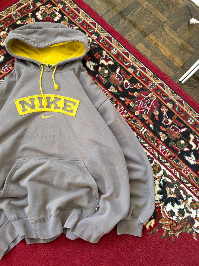 May include: A gray Nike hoodie with a yellow interior hood and yellow drawstring. The Nike logo is prominently displayed in yellow across the chest. The hoodie has a front pocket and is displayed on a red rug.