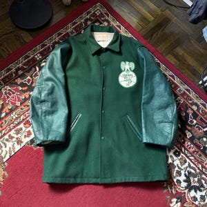 May include: A green wool jacket with dark green leather sleeves. The jacket features a button-up front, two pockets, and a chest patch reading "CHAMPS SS 73". The jacket is displayed on a red rug.