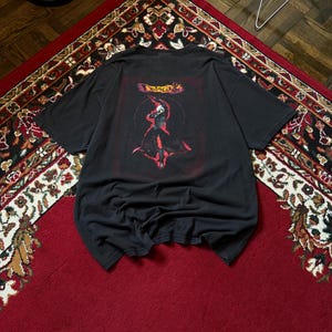 Puede incluir: Camiseta negra con un diseño gráfico en la espalda. El diseño presenta una figura en rojo y negro con la palabra "Dreamcast" arriba. La camiseta está colocada sobre una alfombra roja y estampada.