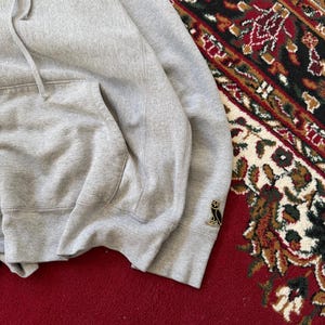 Pu&ograve; includere: Una felpa con cappuccio grigio m&eacute;lange con tasca a marsupio e cappuccio con coulisse. La felpa con cappuccio presenta un piccolo logo di gufo sulla manica. Il capo &egrave; esposto su un tappeto rosso e fantasia.