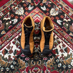 Op de afbeelding: Een paar bruin en zwarte Jordan 13 sneakers. De schoenen hebben een bruin su&egrave;de bovenwerk met zwarte accenten op de neus en veters. De zolen zijn zwart en het Jumpman-logo is zichtbaar op de tong.