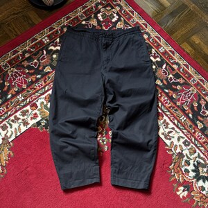 May include: Dark navy cropped trousers with a loose fit. The trousers are made of a solid, dark material and are displayed on a patterned red rug. The trousers have a button and zip fastening.