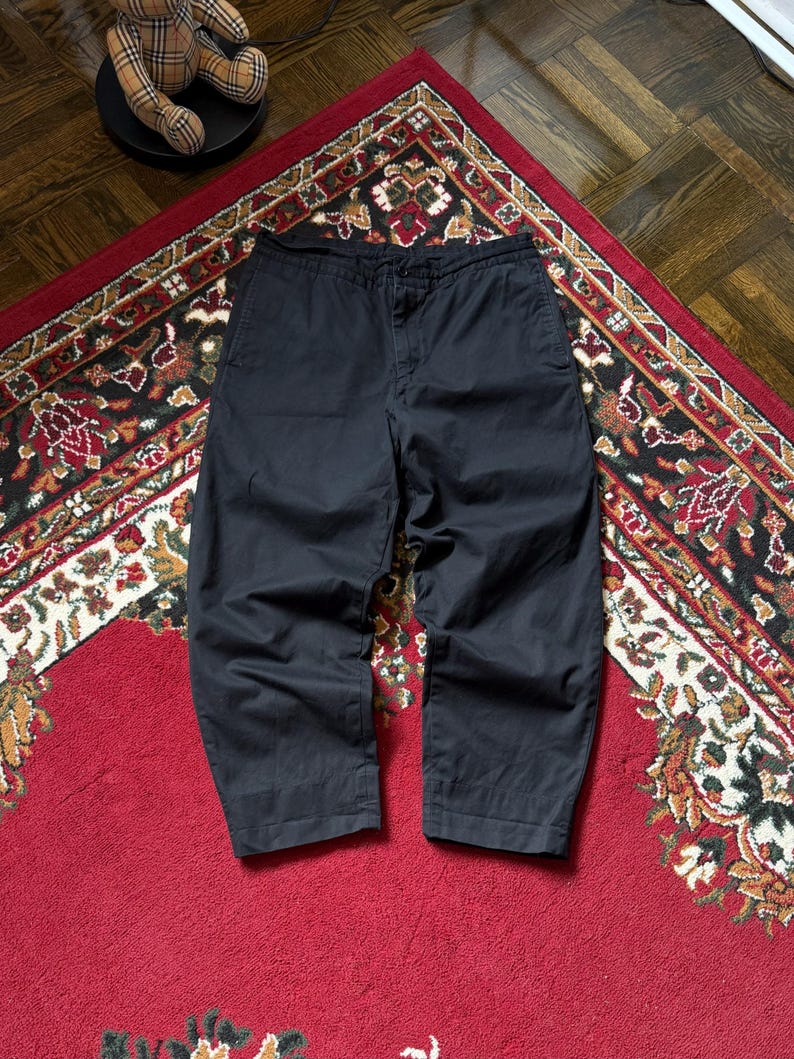 May include: Dark navy cropped trousers with a loose fit. The trousers are made of a solid, dark material and are displayed on a patterned red rug. The trousers have a button and zip fastening.