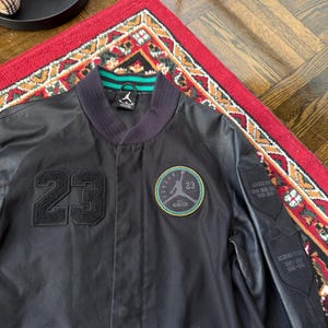 Może przedstawiać: Czarna kurtka bomberka z numerem 23 z przodu. Kurtka ma okrągłą naszywkę z logo i numerem 23. Rękawy mają wygląd sk&oacute;ry. Kołnierz jest granatowy z zielono-białymi paskami.