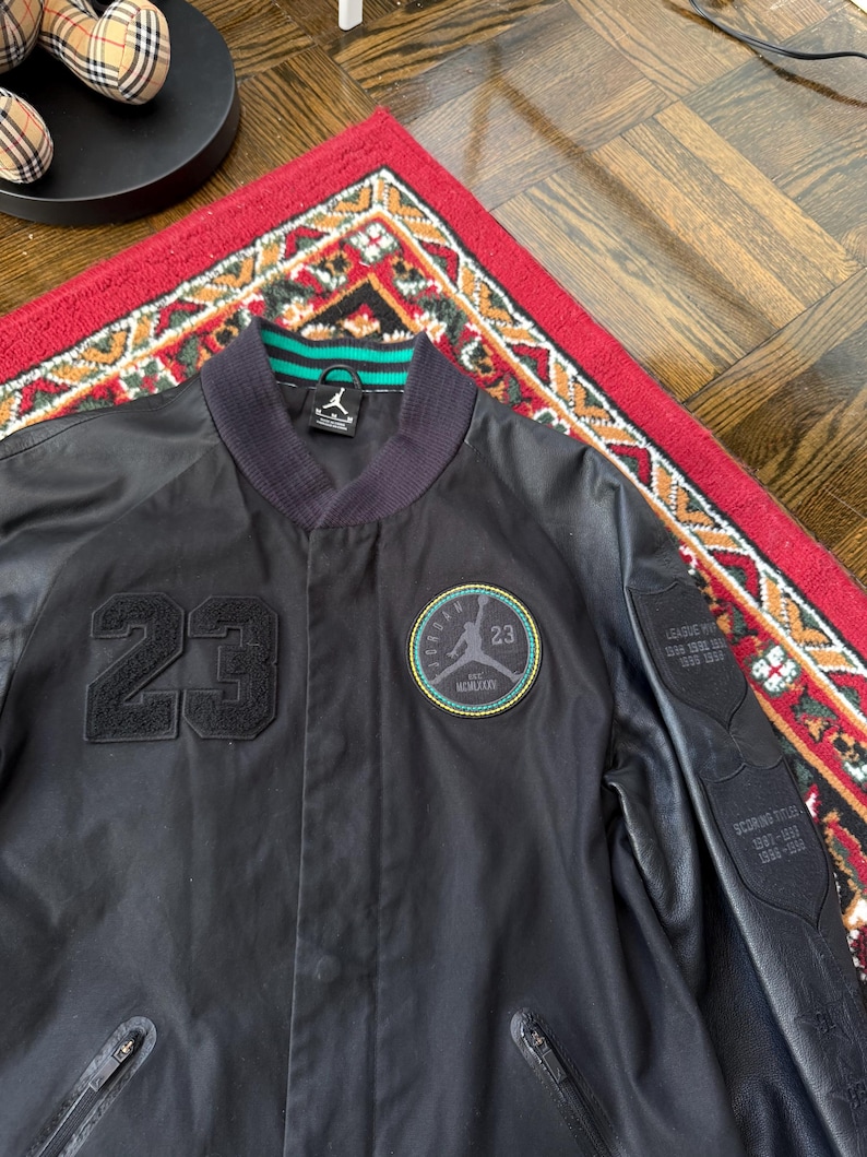 Może przedstawiać: Czarna kurtka bomberka z numerem 23 z przodu. Kurtka ma okrągłą naszywkę z logo i numerem 23. Rękawy mają wygląd sk&oacute;ry. Kołnierz jest granatowy z zielono-białymi paskami.