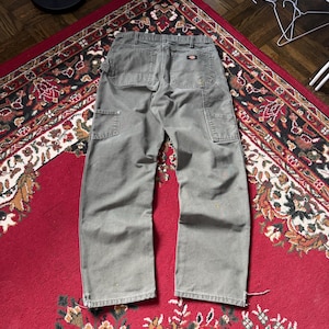 May include: A pair of olive green work pants with multiple pockets, including two side pockets and two back pockets. The pants are laid out on a red patterned rug. The Dickies logo is visible on the back pocket.