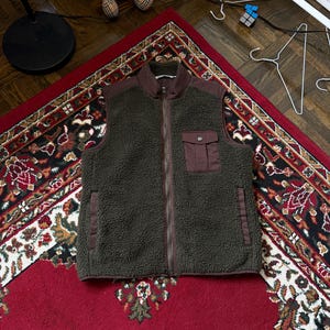 以下が含まれることがあります： フルジップ開閉と茶色の襟とポケットのディテールが特徴の、濃い緑色のフリースベスト。 ベストには胸ポケットが1つあり、模様入りの赤いラグの上に表示されています。