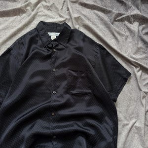 May include: A black short-sleeved button-down shirt with a subtle polka dot pattern. The shirt has a collared neckline, a chest pocket, and a classic button-up design. The fabric has a slight sheen.