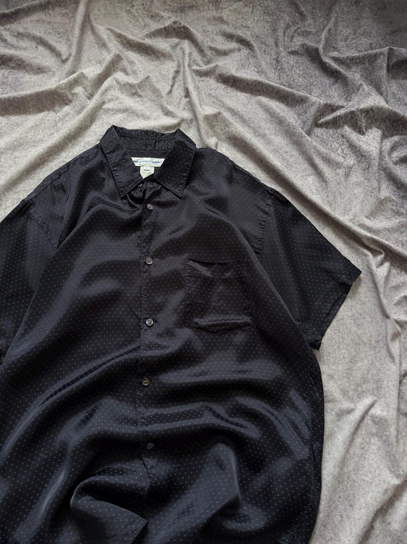 May include: A black short-sleeved button-down shirt with a subtle polka dot pattern. The shirt has a collared neckline, a chest pocket, and a classic button-up design. The fabric has a slight sheen.
