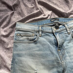 May include: Light blue denim jeans with a faded wash and distressed details. The jeans have a classic five-pocket design, a button and zipper closure, and a brand label at the waistband. The jeans are size 29.