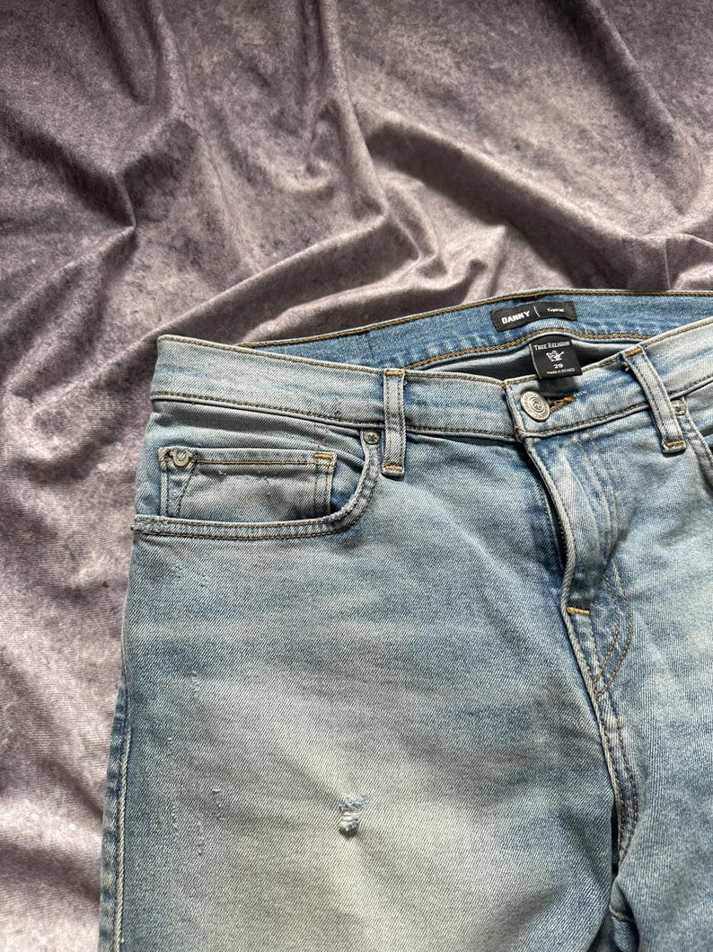 May include: Light blue denim jeans with a faded wash and distressed details. The jeans have a classic five-pocket design, a button and zipper closure, and a brand label at the waistband. The jeans are size 29.