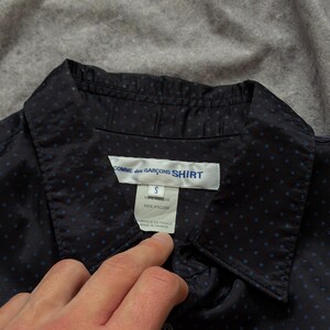 May include: Navy blue button-down shirt with small blue polka dots. The shirt has a collar and a label that reads "COMME des GAR&Ccedil;ONS SHIRT", size "S", and "100% Viscose". The tag also says "FABRIQUE EN FRANCE MADE IN FRANCE".