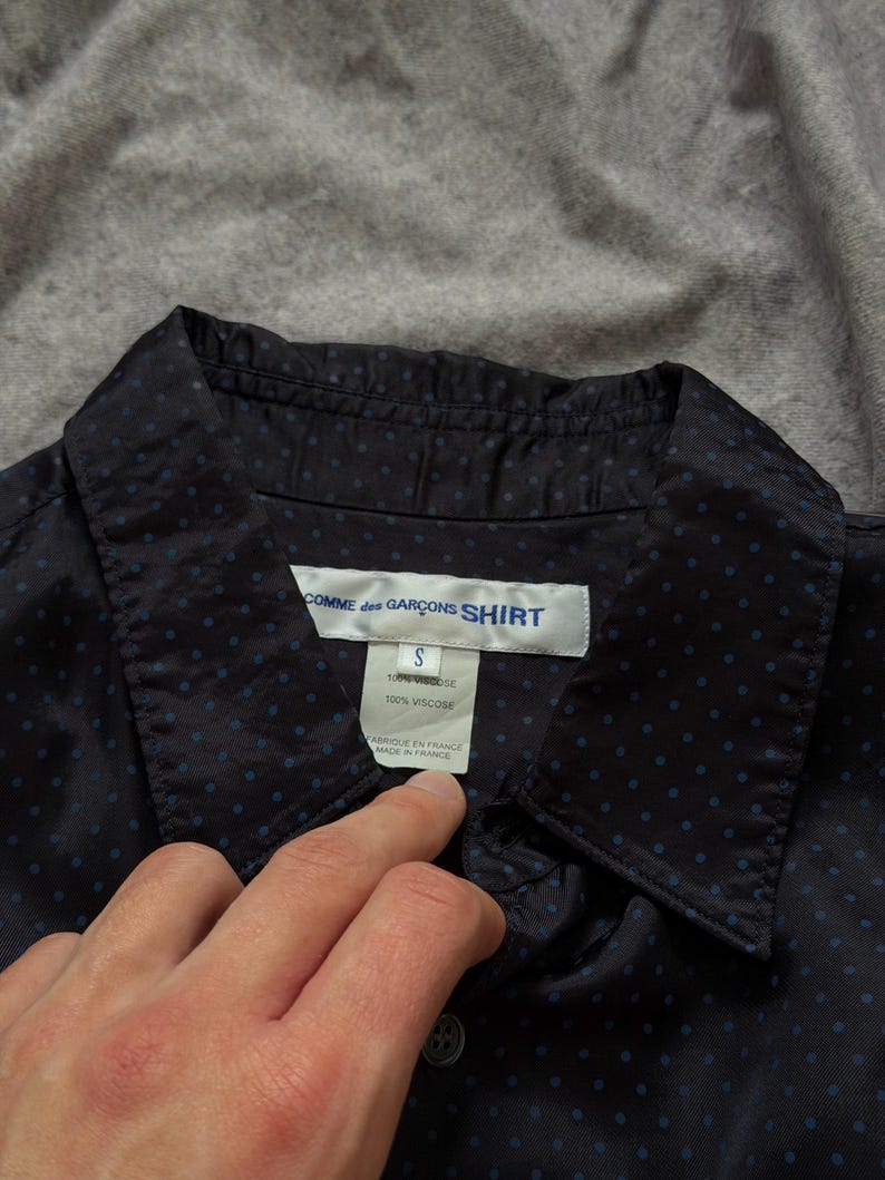 May include: Navy blue button-down shirt with small blue polka dots. The shirt has a collar and a label that reads "COMME des GAR&Ccedil;ONS SHIRT", size "S", and "100% Viscose". The tag also says "FABRIQUE EN FRANCE MADE IN FRANCE".