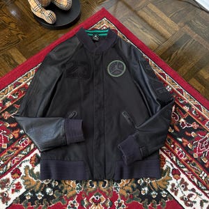 Może przedstawiać: Czarna sk&oacute;rzana kurtka bomberka z numerem "23" z przodu. Posiada zielono-czarną okrągłą naszywkę i zielony pasek na kołnierzu. Kurtka ma prążkowane mankiety i prążkowany d&oacute;ł. Kurtka jest prezentowana na czerwonym dywanie ze wzorem.