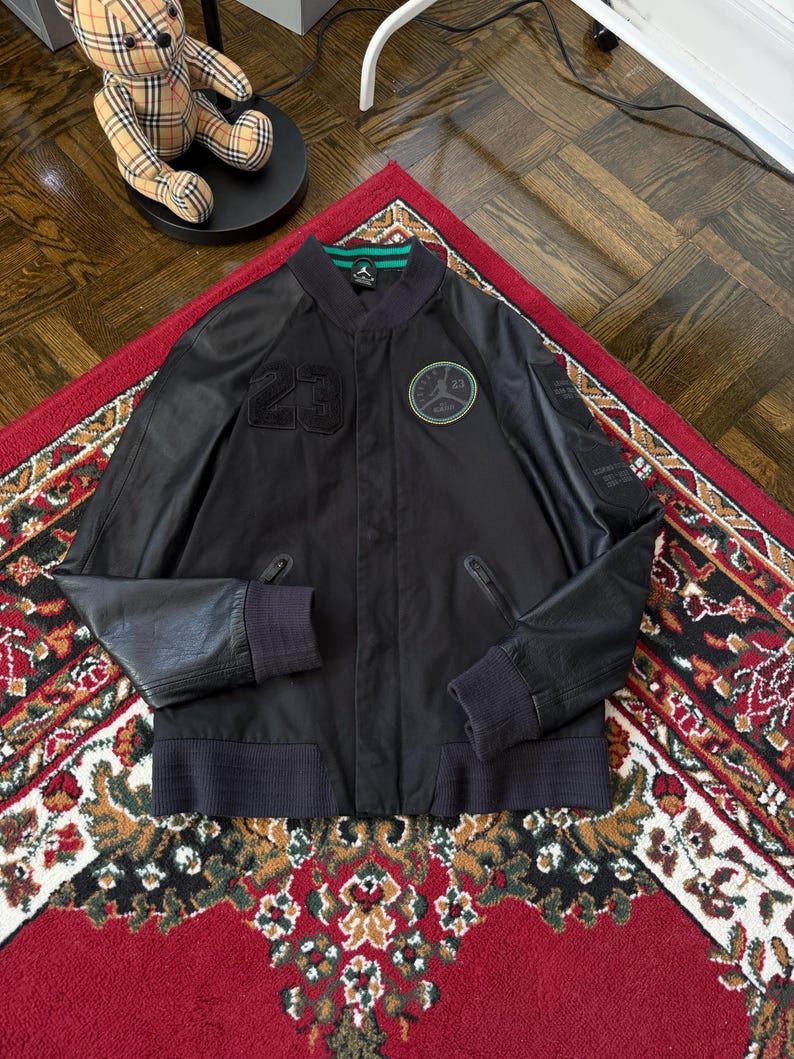 Może przedstawiać: Czarna sk&oacute;rzana kurtka bomberka z numerem "23" z przodu. Posiada zielono-czarną okrągłą naszywkę i zielony pasek na kołnierzu. Kurtka ma prążkowane mankiety i prążkowany d&oacute;ł. Kurtka jest prezentowana na czerwonym dywanie ze wzorem.
