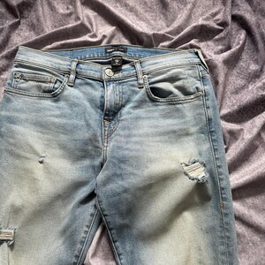 May include: Light blue denim jeans with distressed details. The jeans feature a classic five-pocket design, a button and zipper closure, and a faded wash. The jeans have ripped details on the knees and thighs.