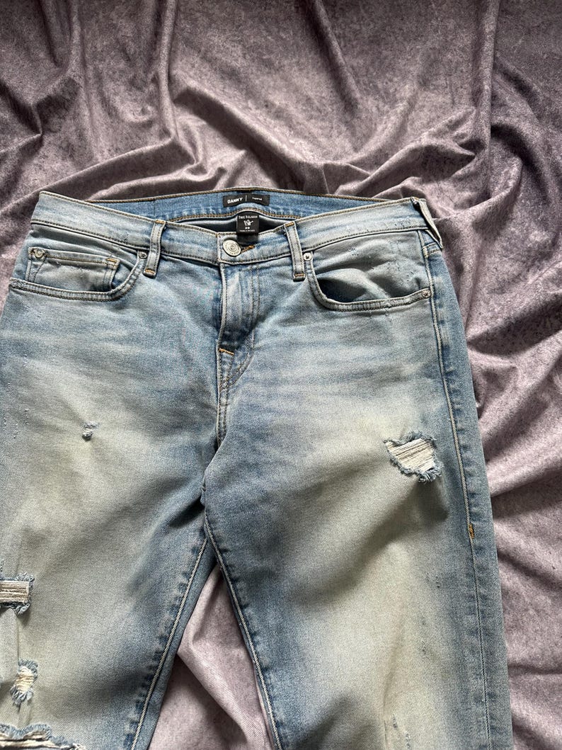 May include: Light blue denim jeans with distressed details. The jeans feature a classic five-pocket design, a button and zipper closure, and a faded wash. The jeans have ripped details on the knees and thighs.