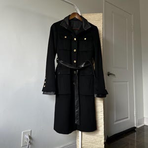 May include: A black coat with a leather collar and belt, hanging on a wooden hanger. The coat features gold buttons and leather accents on the cuffs. The coat is displayed on a white rack.