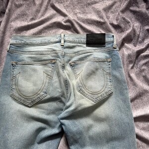 May include: Light blue denim jeans with faded areas and a distressed look. The back pockets feature a stitched design. A black leather patch with white text is visible at the waistband.