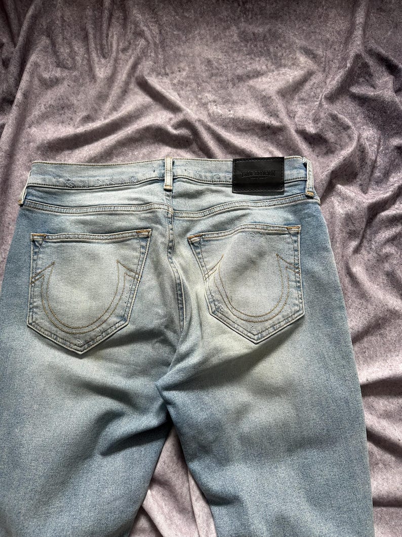 May include: Light blue denim jeans with faded areas and a distressed look. The back pockets feature a stitched design. A black leather patch with white text is visible at the waistband.