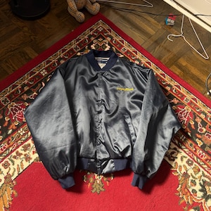 Puede incluir: Chaqueta bomber de satén azul marino con cuello y puños azul oscuro. La chaqueta tiene la palabra "GOODYEAR" bordada en amarillo. La chaqueta se muestra sobre una alfombra roja.