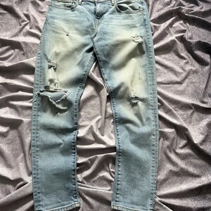 May include: Light blue denim jeans with distressed details, including rips and faded areas. The jeans have a classic five-pocket design and a button and zipper closure. The fabric appears to be a medium-weight denim.
