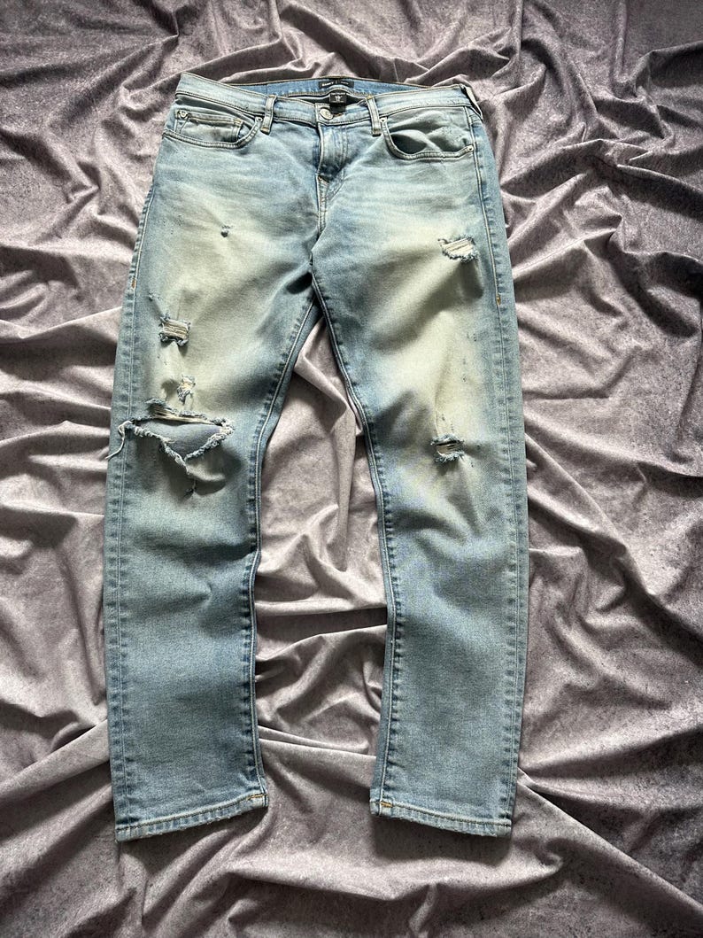 May include: Light blue denim jeans with distressed details, including rips and faded areas. The jeans have a classic five-pocket design and a button and zipper closure. The fabric appears to be a medium-weight denim.