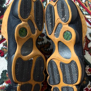 Op de afbeelding: Paar zwarte en bruine sneakers met een textuurzool. De zolen zijn bruin met zwarte rubberen profielen. De schoenen zijn versleten.