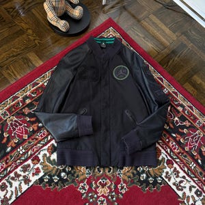 Może przedstawiać: Czarna kurtka bomberka ze sk&oacute;rzanymi rękawami, z numerem 23 i okrągłym logo. Kurtka ma zielono-czarny kołnierz i prążkowane mankiety. Kurtka jest prezentowana na czerwonym dywanie ze wzorem.