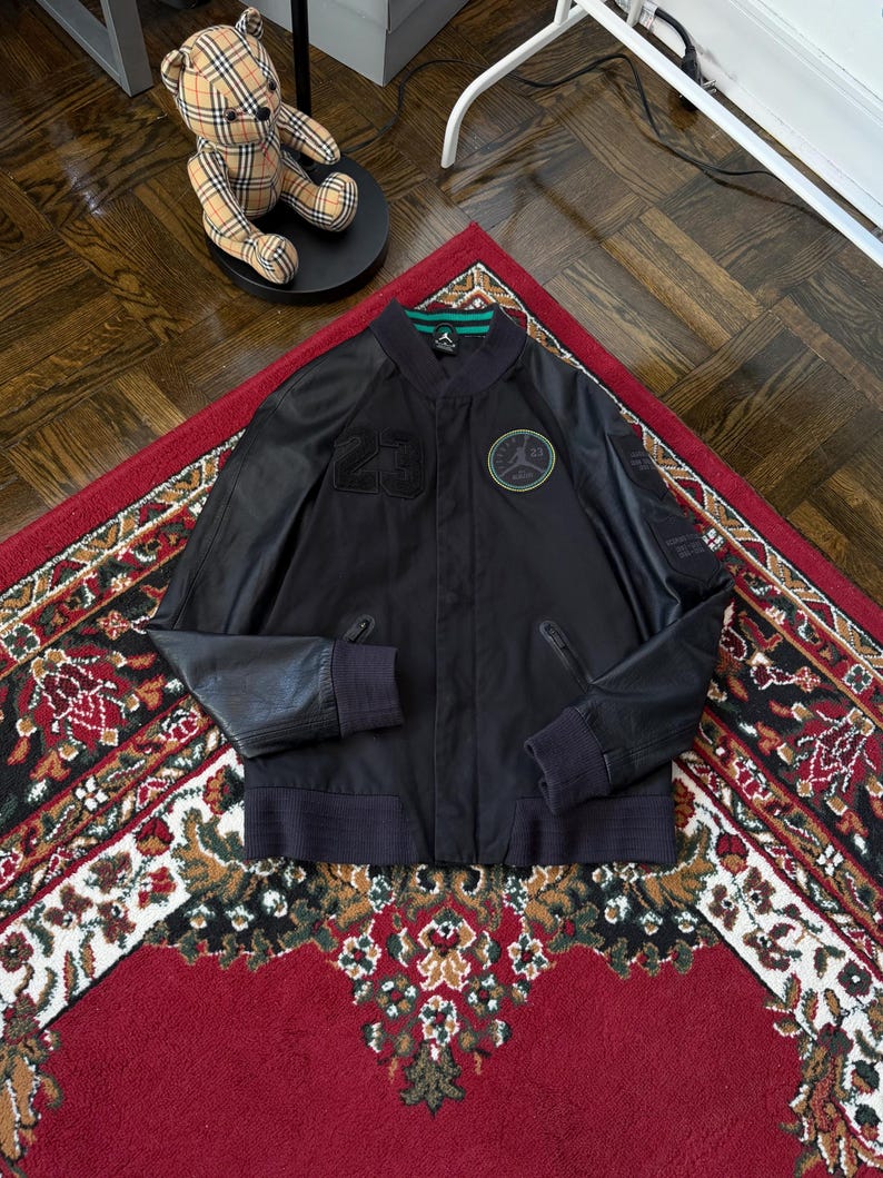 Może przedstawiać: Czarna kurtka bomberka ze sk&oacute;rzanymi rękawami, z numerem 23 i okrągłym logo. Kurtka ma zielono-czarny kołnierz i prążkowane mankiety. Kurtka jest prezentowana na czerwonym dywanie ze wzorem.