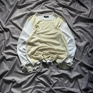 Peut inclure: Un haut à manches longues avec un panneau avant texturé de couleur crème et des manches blanches. Le haut a un col rond et des détails à volants aux poignets. Le vêtement est présenté sur un fond de tissu gris.