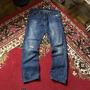 May include: Blue denim jeans with distressed details, including rips at the knees. The jeans are laid flat on a red patterned rug, showcasing their faded wash and classic five-pocket design. The jeans have a button and zipper closure.