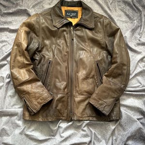 Puede incluir: Chaqueta de cuero marrón con cremallera, dos bolsillos delanteros y cuello. La chaqueta tiene un aspecto desgastado y vintage. El logo de la marca "AJ Armani Jeans" es visible en el cuello.